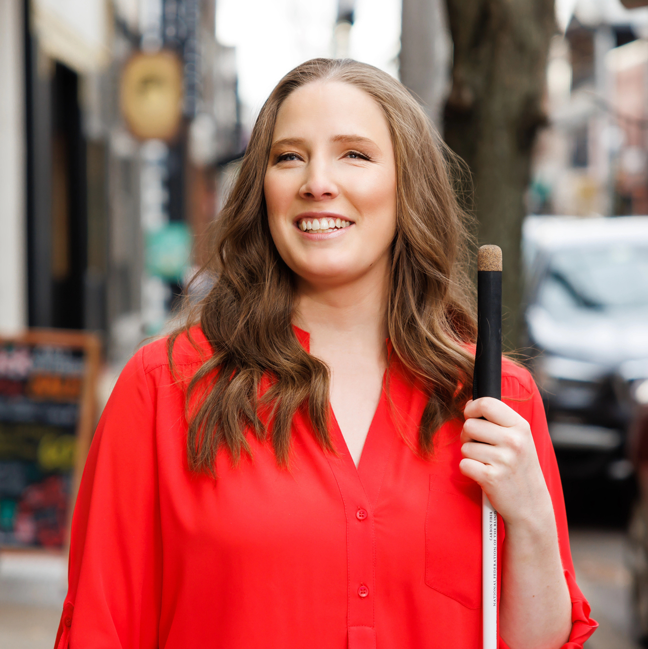 A portrait of Cynthia smiling slightly at three-quarters view and gazing upwards. She is seen from the elbows up, wearing a bright crimson blouse and holding her white cane. Behind her are hints of a bustling Pittsburgh, PA street.