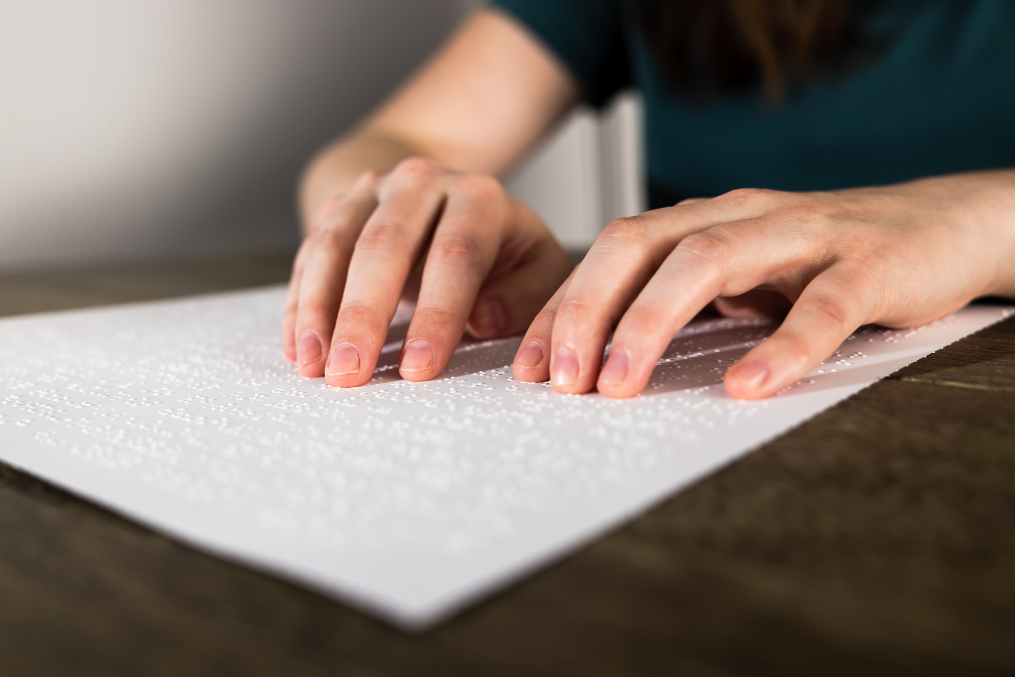 Cynthia’s hands shot from the front, reading a page of braille writing (raised tactile markings). The page rests on a dark wooden table.