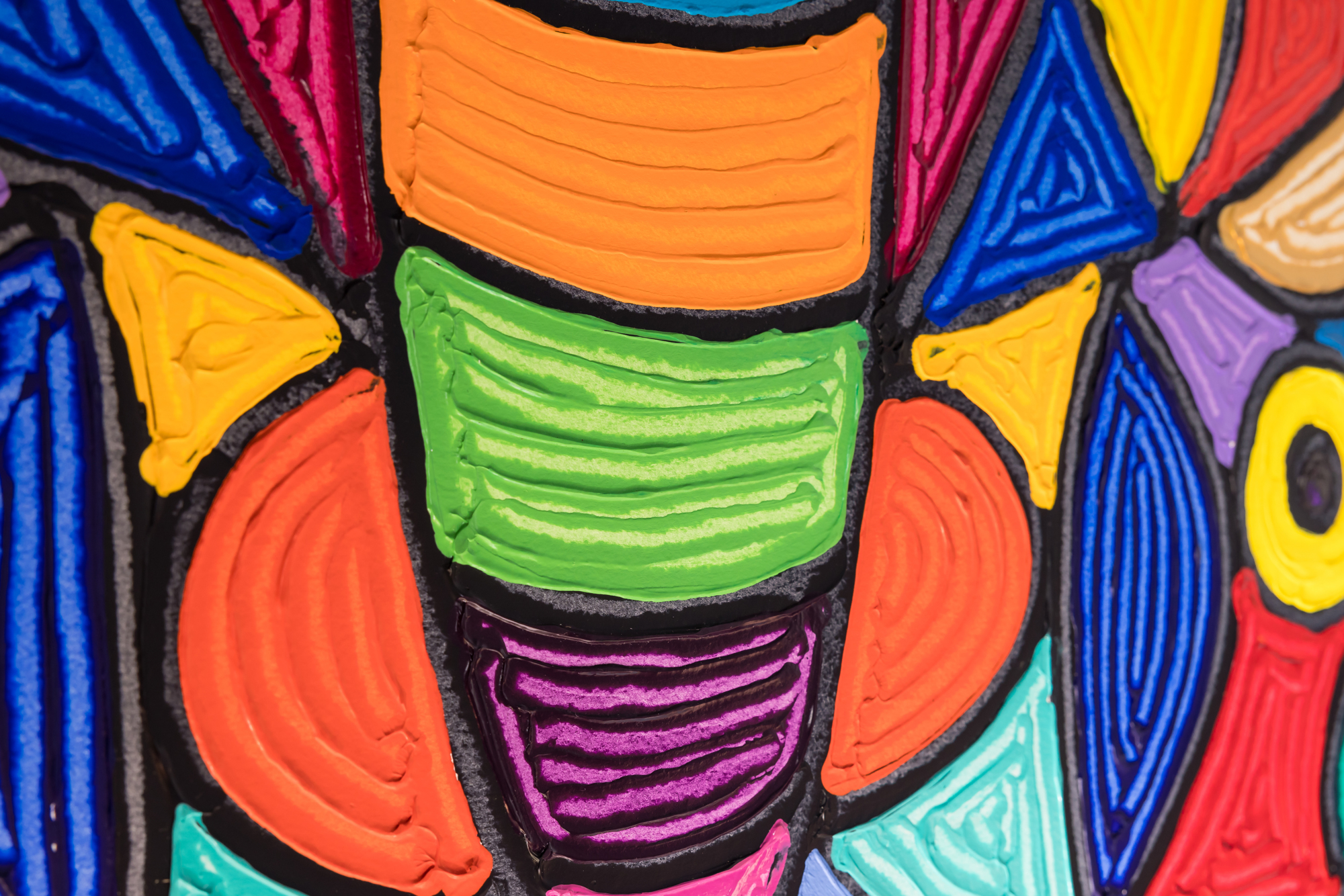 A closeup of a large, abstract butterfly painting. The butterfly is rendered in bright blocks of color, using raised paint strokes that suggest a finger painting technique. The photograph zooms in on the butterfly’s body, rendered in bold horizontal stripes of color in orange, green, and purple with black outlines. On either side of the butterfly body, there are symmetrical colored shapes that make up the wings, like dark orange semi-circles and blue, yellow, and red triangles.