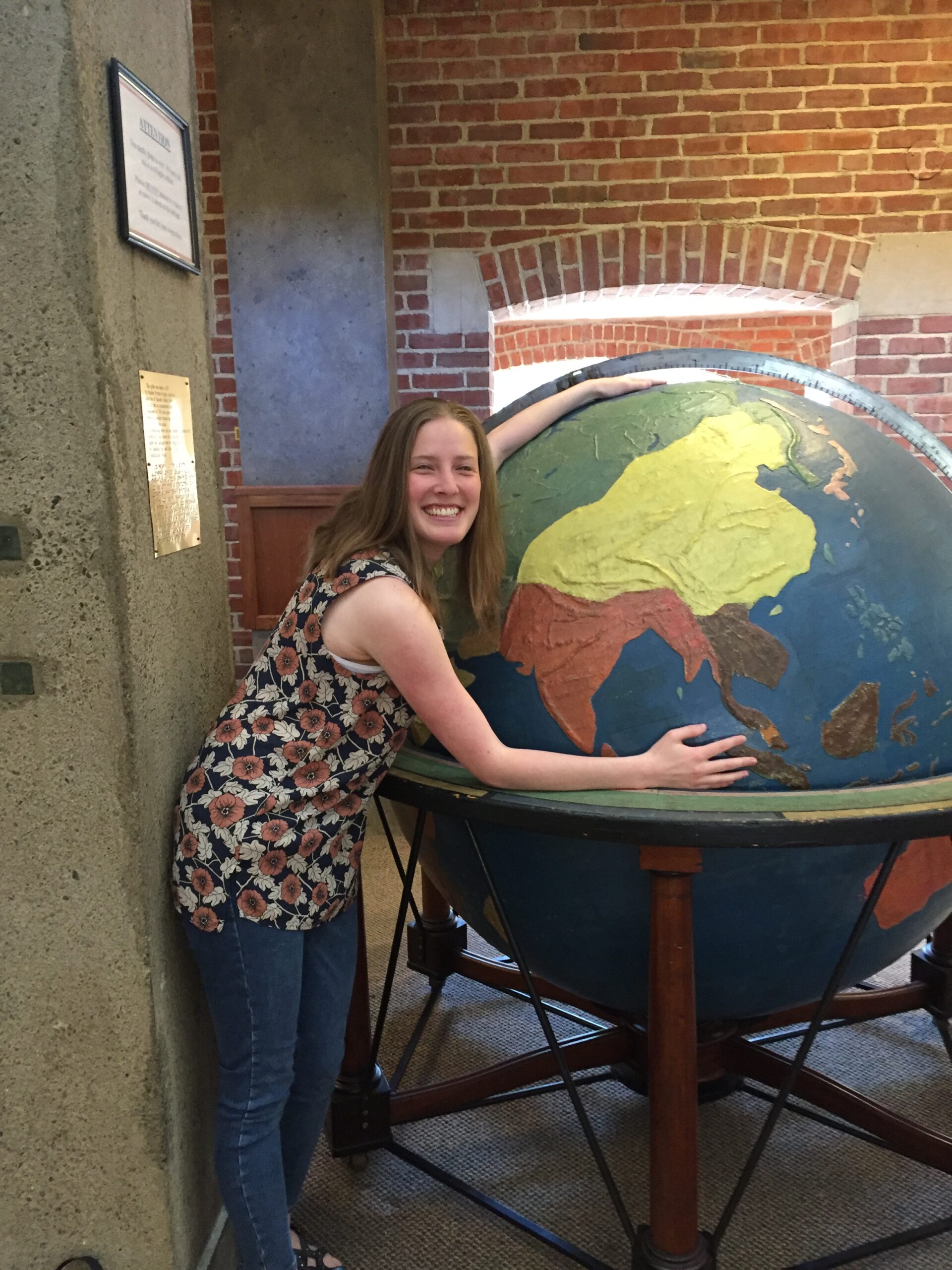 Cynthia is pictured in full length with her arms reaching around a giant, tactile globe at the Perkins School for the Blind. The globe is nearly her height, and is turned to show regions of Southeast Asia and Australia in yellow, brown, and green. Cynthia is grinning, and wears a flower print blouse and blue jeans.